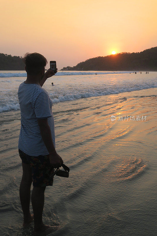 20岁出头的亚洲印度游客在海滩上自拍，手持手机，自拍男子在度假果阿海滩用手机自拍日落，t恤，沙子，海边日落/日出的剪影